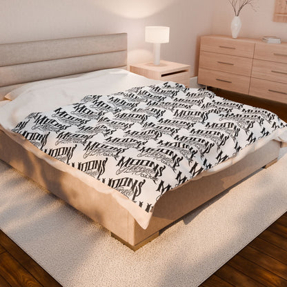 MOONSMC OG Logo plush blanket spread across a bed in a softly lit bedroom.