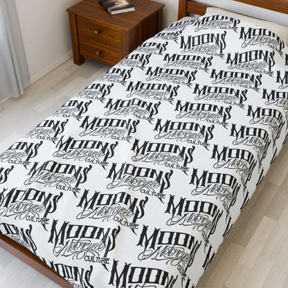 MOONSMC OG Logo plush blanket displayed lengthwise on a bed beside a wooden nightstand.