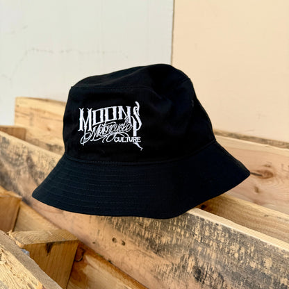 MOONSMC OG Logo Embroidered Bucket Hat: black embroidered bucket hat resting on a wooden railing.