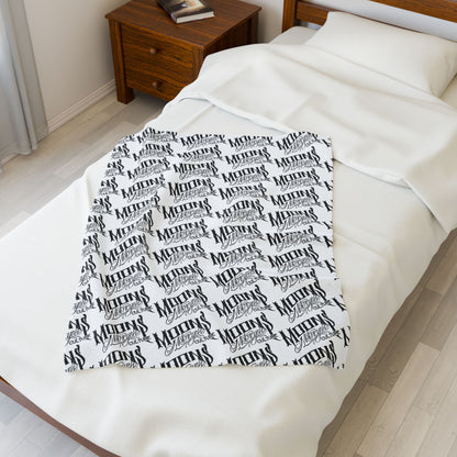 MOONSMC OG Logo plush blanket displayed lengthwise on a bed beside a wooden nightstand.
