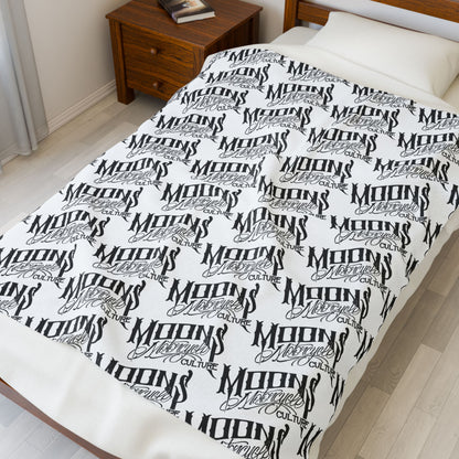 MOONSMC OG Logo plush blanket displayed lengthwise on a bed beside a wooden nightstand.