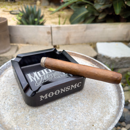 MOONSMC "The Diplomat" Cigar Ashtray: black tray holding a cigar on an outdoor table.